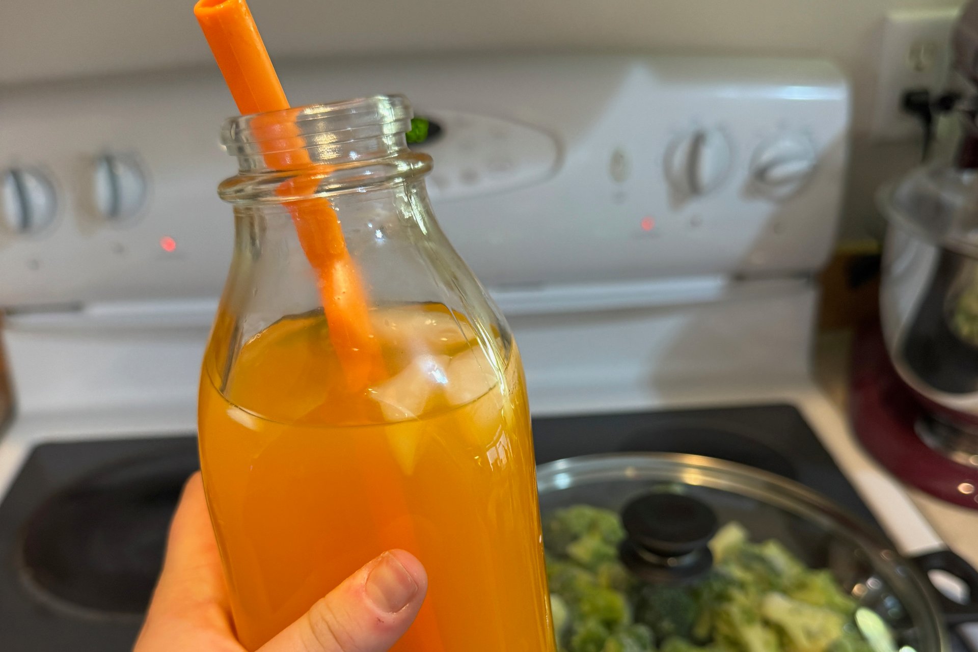 Hand holding a glass bottle of orange hydration drink with ice and an orange straw in a home kitchen.